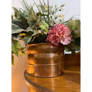 May include: A cylindrical copper planter with a polished metallic finish and horizontal ridges, containing an arrangement of artificial greenery, ivy leaves, small white flowers, and a large, ruffled pink peony bloom.