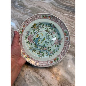 May include: A round ceramic plate with a light green background featuring a central illustration of a blue bird perched on a branch surrounded by pink and yellow flowers. The rim has a geometric red lattice pattern with small floral accents.