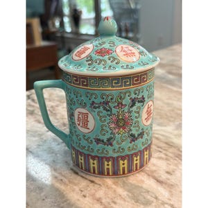 May include: A light blue ceramic mug with a matching lid, featuring a colorful floral pattern, intricate scrollwork, and red Chinese characters. The design includes a geometric border near the rim and a vertical striped pattern at the base.