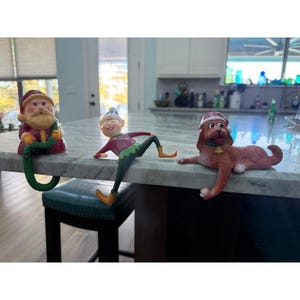 May include: Three shelf-sitting figurines including a Santa Claus in a red suit holding a green rope, an elf in a red and green outfit with a pointed hat, and a brown dog wearing a red Santa hat and a green collar with a gold bell.