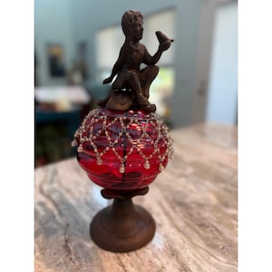 Op de afbeelding: Een decoratieve glazen pot met een dieprood, geribbeld lichaam en een donkerbruine metalen voet. Het deksel is voorzien van een donkerbruine metalen figuur van een persoon die een klein vogeltje vasthoudt. Een fijne, gedrapeerde ketting van kraaltjes.