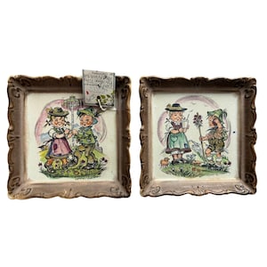 Puede incluir: Dos baldosas de cerámica enmarcadas con ilustraciones coloridas de niños con trajes tradicionales alemanes. Las baldosas tienen un borde decorativo y ornamentado. Las ilustraciones representan a niños en un entorno rural, con texto en la parte inferior de cada baldosa.