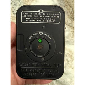 May include: A black rectangular Eastman Kodak film magazine with white text reading: PLACE IN CAMERA THIS SIDE OUT AND WITH THIS END TOWARD LENS, MADE IN ROCHESTER, N. Y., U. S. A. BY EASTMAN KODAK COMPANY, UNEXPOSED FOOTAGE, LOADED WITH KODAK FILM.