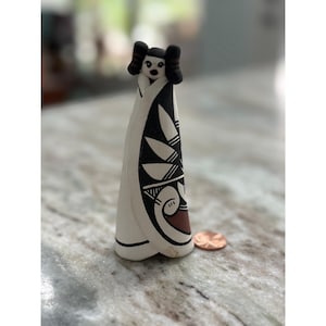 May include: A small, cone-shaped ceramic figurine featuring a white base with black and reddish-brown painted geometric patterns. The figure has a stylized face and dark hair, standing next to a copper penny for scale to show its small size.