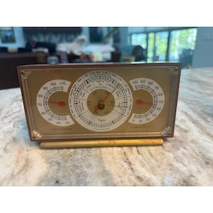 Op de afbeelding: Een rechthoekig Taylor Stormoguide weerstation met een goudkleurige wijzerplaat. Het apparaat heeft drie ronde meters: een temperatuurmeter aan de linkerkant, een centrale barometer met stijgende en dalende indicatoren en een vochtigheidsmeter rechts.