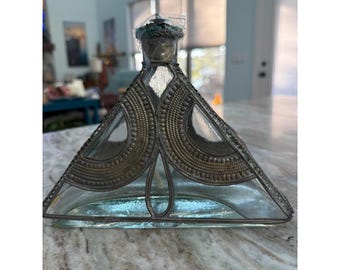 Könnte beinhalten: Eine dreieckige Glasflasche mit einem dekorativen Metallüberzug und komplizierten Perlenmustern. Die Flasche hat einen schmalen Hals, der von einem kleinen, strukturierten Metallverschluss gekrönt wird. Der klare Glaskörper ist von dunklem Filigran.
