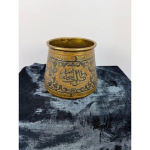 May include: A cylindrical brass vessel featuring intricate dark etched floral patterns and Arabic calligraphy encircling the exterior. The metal surface displays a warm, aged patina with contrasting dark and golden tones throughout the design.