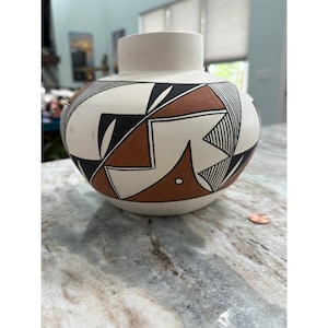 May include: A round ceramic vase with a matte white base featuring geometric patterns in black and terracotta brown. The design includes sharp angular shapes, fine parallel lines, and small circular accents. A copper penny sits nearby for scale.