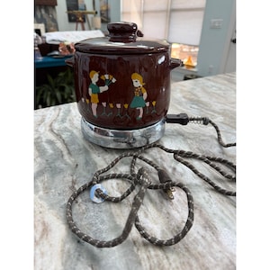 May include: A brown ceramic electric slow cooker with a matching lid, featuring a painted graphic of two figures holding flowers among tulips. The pot sits on a circular chrome base and includes a brown and white fabric-covered electrical power cord.