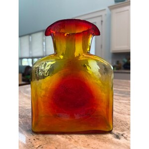 Pode incluir: Um vaso de vidro quadrado com um gradiente de cores vibrantes, transitando de um centro vermelho profundo para um exterior amarelo e laranja brilhante. O recipiente possui uma borda larga e alargada com um acabamento liso e translúcido.
