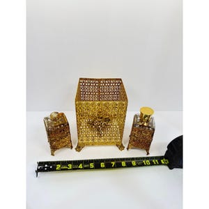 May include: A gold-tone metal vanity set consisting of a tall rectangular open-top container and two smaller matching glass bottles with ornate filigree metal casings. A yellow measuring tape shows the set spans approximately 11 inches wide.