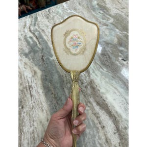 May include: A hand mirror with a gold-toned metal frame and handle. The back of the mirror features a cream-colored fabric insert with a central oval medallion containing an embroidered floral design in shades of pink, blue, and green.