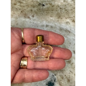 Op de afbeelding: Een kleine, heldere glazen parfumfles in de vorm van een kroon, met een goudkleurige dop. De fles wordt in een hand gehouden, wat het delicate ontwerp en de grootte benadrukt. De kroonvorm voegt een vleugje elegantie toe.