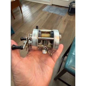 May include: A silver-toned metal fishing reel featuring a dual-handle design with black plastic knobs, a central spool wound with gold-coloured fishing line, and a metallic frame with visible mechanical components and a mounting bracket.