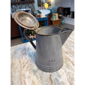 May include: A gray enameled metal pitcher with a textured finish, a hinged lid, a side handle, and a pouring spout. The pitcher shows signs of wear with visible chips in the enamel coating along the rim and body, revealing the metal underneath.