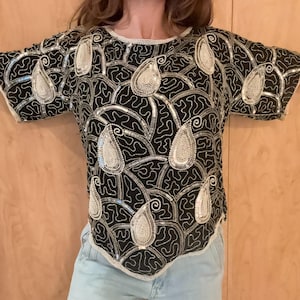 May include: A short-sleeved black top featuring an intricate pattern of silver sequins and white beaded paisley motifs. The garment has a round neckline and a straight hem, with the entire surface covered in dense, reflective beadwork and embroidery.