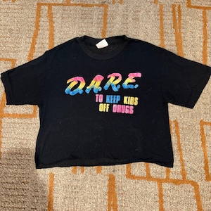 May include: Black t-shirt with the text "DARE TO KEEP KIDS OFF DRUGS" in a colorful, gradient font. The shirt is short-sleeved and has a crew neck. The text is in a rainbow of colors, including blue, yellow, and pink.