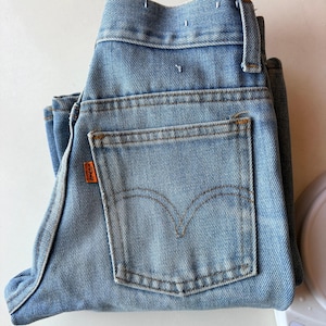 May include: Light blue denim jeans with a classic five-pocket design. The jeans feature a back pocket with a stitched arcuate design and an orange Levi's tag. The jeans are folded and appear unworn.
