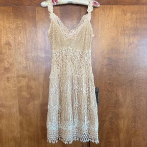 May include: A sleeveless beige dress featuring an intricate lace overlay with a V-neckline. The bodice displays a floral lace pattern, while the skirt section transitions into a vertical, open-work crochet design with a scalloped, fringed hemline.