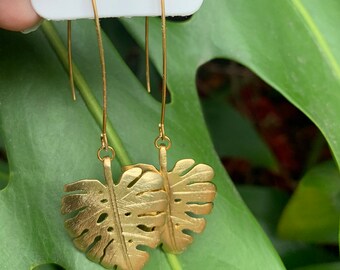 May include: A pair of gold-toned drop earrings featuring a textured monstera leaf design. The earrings are attached to a white display card and hang from long, thin, curved gold-toned wires that extend upward from the leaf-shaped pendants.