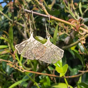 May include: Gold-coloured earrings featuring a ginkgo leaf design. The earrings hang from thin wires, set against a backdrop of green foliage and branches. The earrings have a detailed, textured surface, and are a stylish accessory.