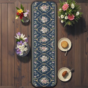 Op de afbeelding: Een marineblauwe loper met een bloemenpatroon van roze en witte rozen. De loper heeft een gouden rand en ligt op houten planken. Twee kleine borden met eten en bloemen in vazen zijn ook aanwezig.
