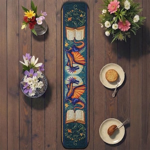May include: A teal table runner with a dragon design. The runner features a repeating pattern of dragons with orange wings, open books, and floral accents. The runner is placed on a wooden table with flowers and plates.