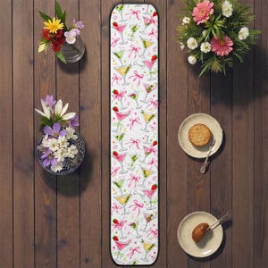 Op de afbeelding: Een witte tafelloper met een cocktailontwerp. De loper heeft martini glazen in roze, groen en geel, versierd met roze strikken en aardbeien. De loper is omgeven door bloemen en kleine bordjes met eten.