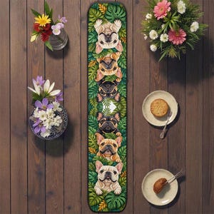May include: A long, rectangular table runner featuring a repeating pattern of French bulldog illustrations set against a backdrop of green tropical leaves. The dogs are in various colors, including tan, black, and white. The runner is placed on a wooden surface.