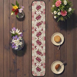 May include: A floral table runner with a cream background and repeating burgundy rose pattern. The runner is surrounded by decorative elements, including vases of flowers and plates with pastries, all arranged on a wooden surface.