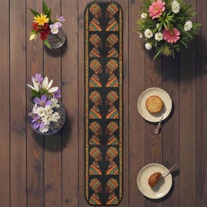 以下が含まれることがあります： カラフルなヘッドラップをかぶったアフリカ人女性の繰り返し模様が特徴の黒いテーブルランナー。 ランナーは、花瓶に入った花と、木製のテーブルに置かれた食べ物の入ったお皿に囲まれています。