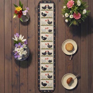 May include: A long, rectangular table runner with a repeating pattern of black and white chickens. The runner is bordered with a dark blue floral design. The image also includes floral arrangements and plates with food.
