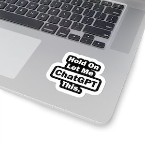 May include: A white sticker with the text "Hold On Let Me ChatGPT This" in black. The sticker is placed on a silver laptop, partially covering the keyboard. The text is in a bold, sans-serif font.