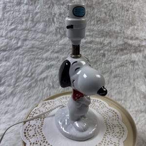 May include: A white Snoopy lamp with black ears and a black nose. Snoopy is wearing a red polka dot bow tie. The lamp has a white base and a white lamp holder.