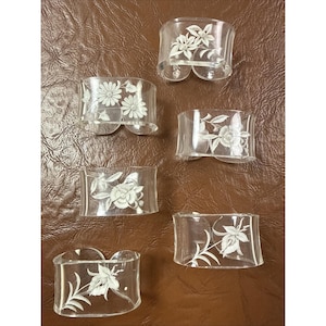 May include: Six clear glass napkin rings with etched floral designs. Each ring features a different floral pattern, including daisies and various stylized flowers. The rings are curved and have a small opening on the bottom.