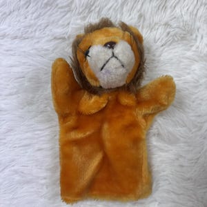 May include: A plush lion hand puppet in shades of orange and brown. The puppet has a white face with a brown nose and whiskers. The lion's mane is a darker brown, and the puppet is made of soft, fuzzy material.