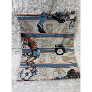 May include: A rectangular fabric pillowcase featuring a vintage North American Soccer League print. The design includes horizontal blue and orange stripes, illustrations of soccer players in action, and various team logos like NASL, Kicks, and Cosmos.