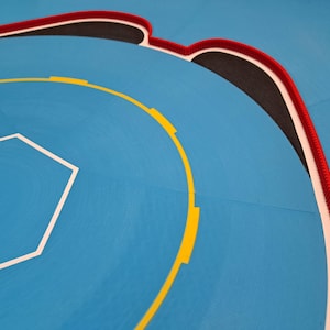 May include: A section of a blue wrestling mat with white, yellow, black, and red markings. The mat features a white hexagonal shape and a curved yellow line with yellow rectangles. A red and white braided border outlines the edge.