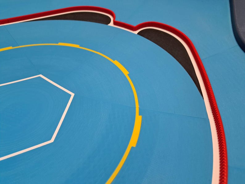 May include: A section of a blue wrestling mat with white, yellow, black, and red markings. The mat features a white hexagonal shape and a curved yellow line with yellow rectangles. A red and white braided border outlines the edge.