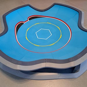 May include: A blue and gray arena with a hexagonal center and concentric circles in yellow, red, and white. The arena has a unique, curved shape with a raised edge, designed for competitive play.