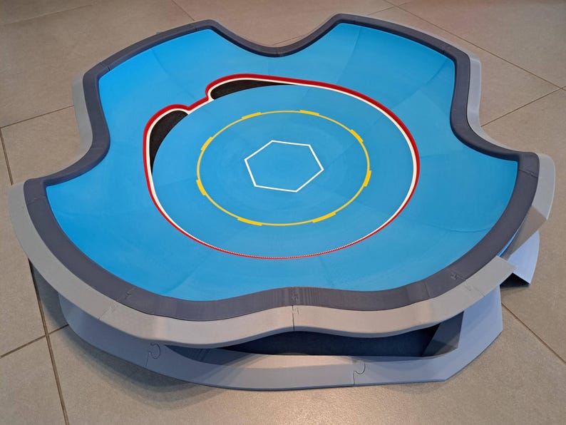 May include: A blue and gray arena with a hexagonal center and concentric circles in yellow, red, and white. The arena has a unique, curved shape with a raised edge, designed for competitive play.