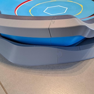 May include: A multi-layered, circular game arena with a blue and gray color scheme. The top layer features a blue surface with a white hexagon and red and yellow accents. The arena has a curved, gray outer edge and a blue interior.