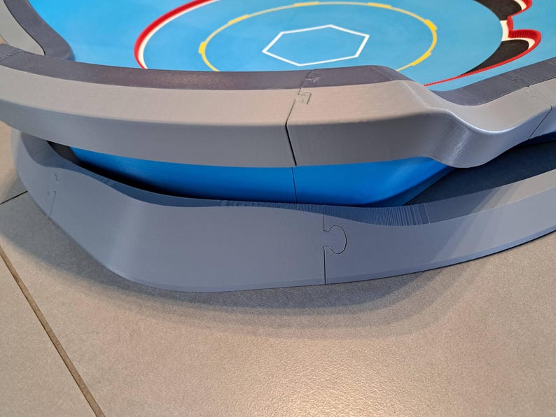 May include: A multi-layered, circular game arena with a blue and gray color scheme. The top layer features a blue surface with a white hexagon and red and yellow accents. The arena has a curved, gray outer edge and a blue interior.