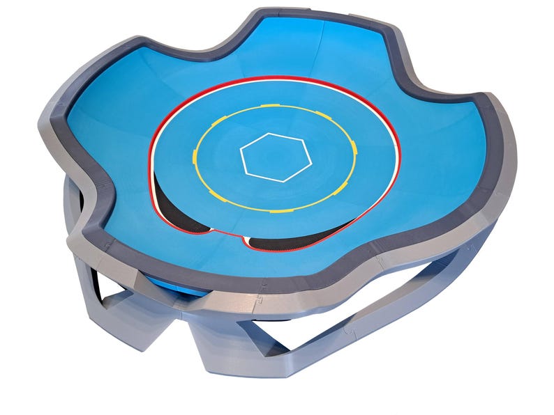 May include: A blue and gray wrestling ring with a unique, wavy shape. The ring features a light blue mat with a red, yellow, and white circular design. The ring is supported by a gray frame.