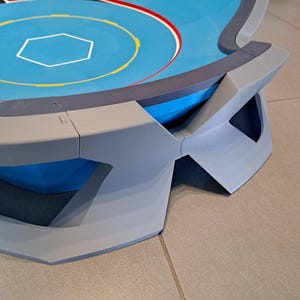 May include: A blue and gray arena with a hexagonal center and concentric circles. The arena has a raised edge with geometric supports. The surface is blue with yellow, red, and white markings. The design is modern and angular.