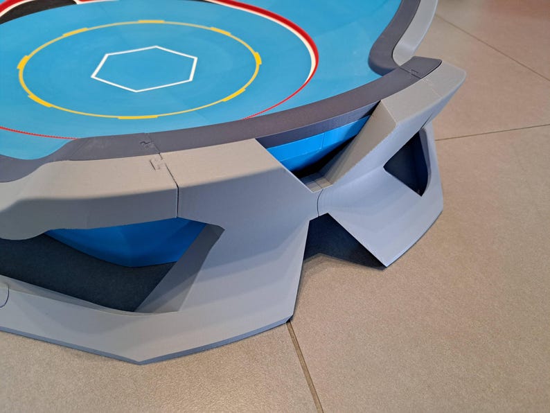 May include: A blue and gray arena with a hexagonal center and concentric circles. The arena has a raised edge with geometric supports. The surface is blue with yellow, red, and white markings. The design is modern and angular.
