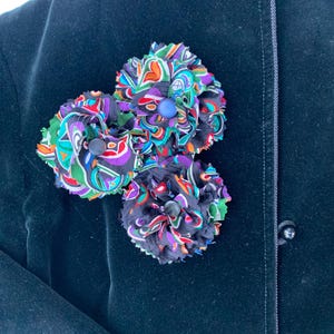 May include: Three fabric flower brooches in vibrant colors of green, orange, purple, and black are pinned to a dark navy blue velvet jacket. Each flower has a button center. The jacket has a tailored design with button closures.