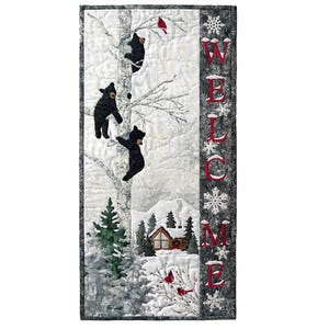May include: A rectangular quilted wall hanging featuring a winter scene with three black bears climbing a white birch tree, a small cabin, and a red cardinal. The word WELCOME is vertically embroidered in red letters on a dark grey border.