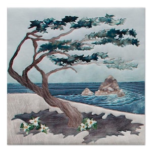 May include: A square textile art piece featuring a windswept tree with a brown trunk and dark teal foliage. The scene includes a blue ocean with rocky formations, a light gray sandy foreground, and small white flowers with green leaves at the base.