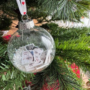 May include: A clear glass Christmas ornament filled with small, square photos and white confetti, hanging from a white ribbon with red heart accents. The ornament is suspended among green Christmas tree branches.
